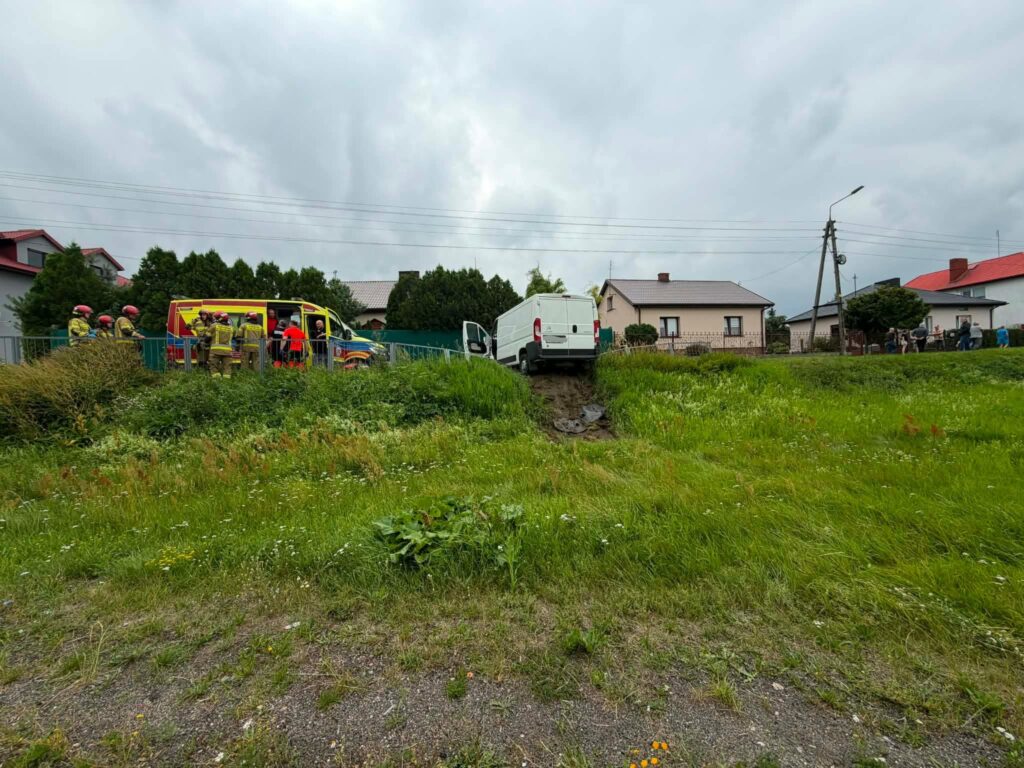 Grupa strażaków w hełmach pomaga przy wypadku, obok stoi ambulans i biały van na mokrej ziemi w zielonym terenie.