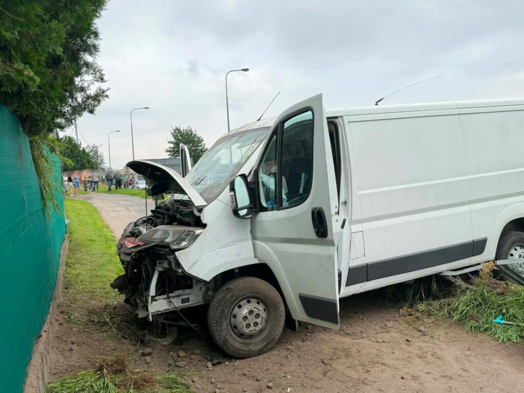 Biały furgon z uszkodzoną przednią częścią stoi na trawie obok drogi, z otwartą maską i drzwiami. W tle widać grupę ludzi.