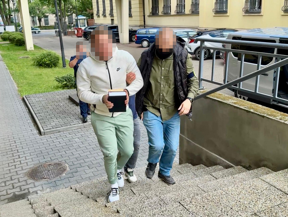 Mężczyzna w jasnym dresie prowadzony przez funkcjonariusza policji w kierunku schodów w miejskim otoczeniu.
