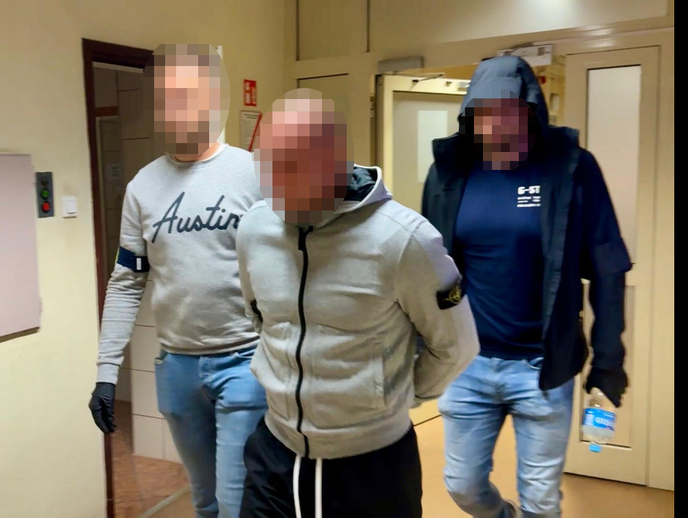 Mężczyzna w szarym dresie prowadzony przez dwóch funkcjonariuszy w korytarzu budynku.