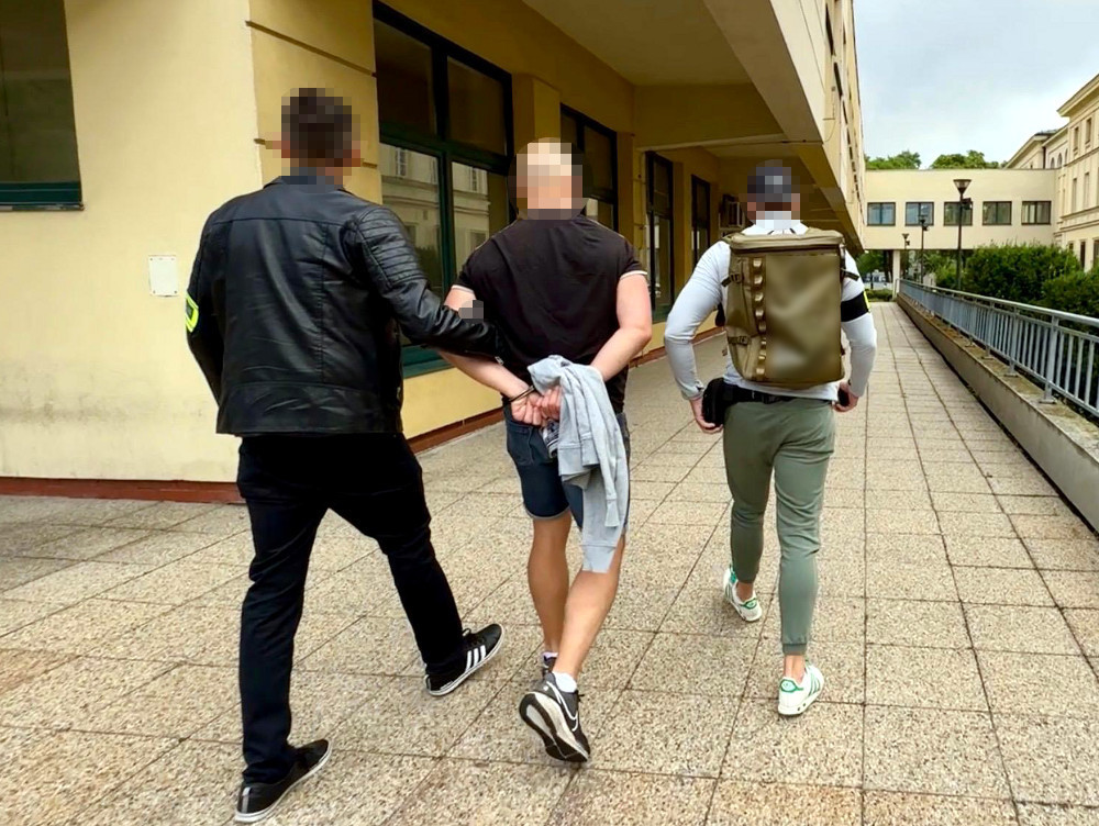 Dwaj mężczyźni prowadzą innego mężczyznę, który ma związane ręce, wzdłuż chodnika w budynku.