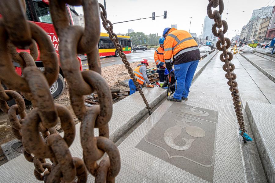 Pracownicy w pomarańczowych kamizelkach roboczych montują płyty na torowisku podczas remontu w miejskim otoczeniu.