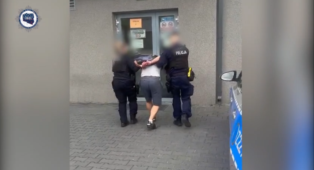 Mężczyzna w szortach prowadzony przez dwóch policjantów w mundurach w pobliżu budynku z szarym tłem.