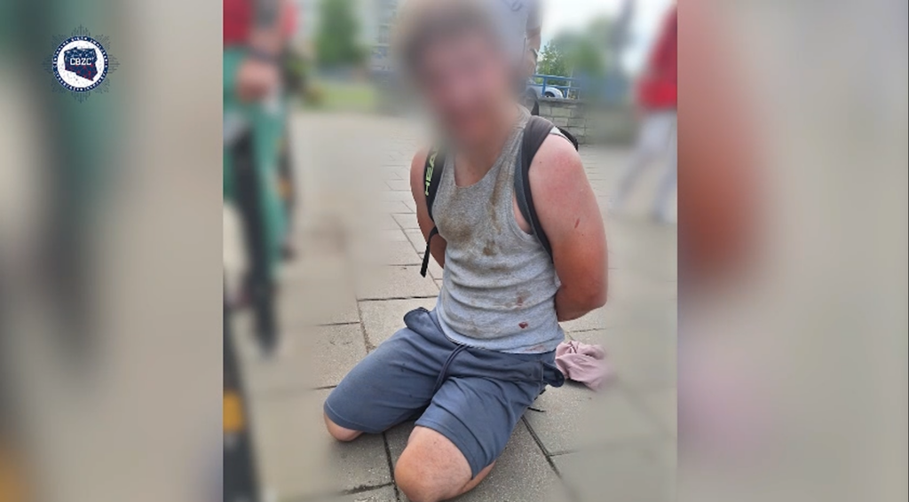Mężczyzna w szarej koszulce klęczy na chodniku, z rękami związanymi za plecami, w otoczeniu przechodniów.