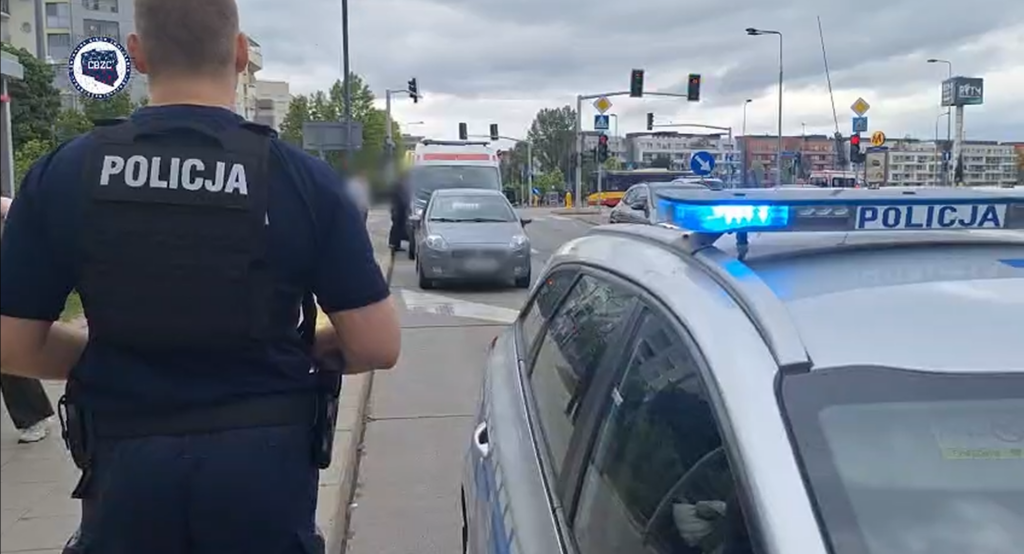 Policjant w kamizelce z napisem "POLICJA" stoi obok radiowozu z włączonymi sygnałami świetlnymi na ruchliwej ulicy.