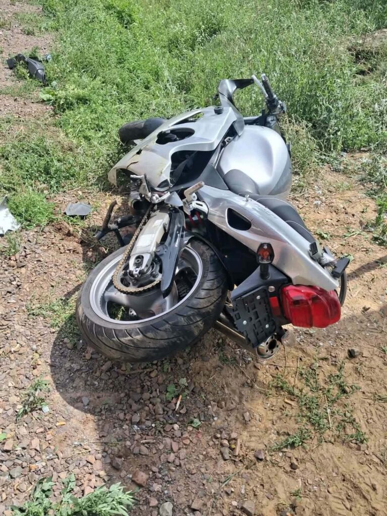 Zniszczony motocykl leży na boku na trawie obok drogi, otoczony kamieniami i zielenią.