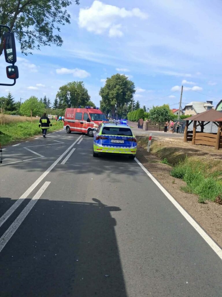 Na drodze stoi radiowóz policyjny oraz karetka pogotowia, obok przechodzą ludzie w tle.