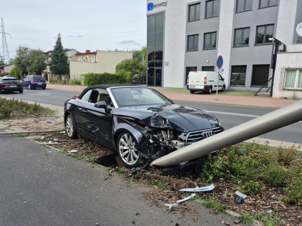 Czarny samochód sportowy uderzył w stalowy słup, znajdując się na poboczu drogi w miejskim otoczeniu.