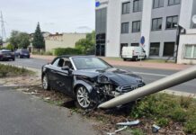 Czarny samochód sportowy uderzył w stalowy słup, znajdując się na poboczu drogi w miejskim otoczeniu.