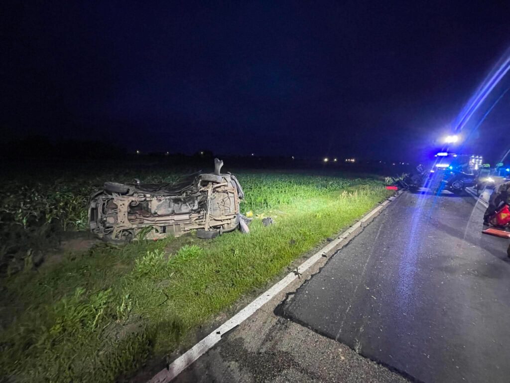 Przewrócony samochód osobowy leży na poboczu drogi, obok rosnącego zboża, w tle widać policyjne światła.