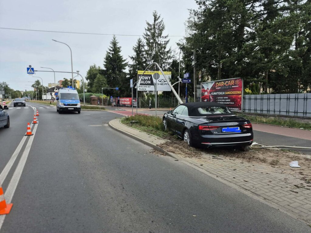 Policyjny radiowóz z włączonymi sygnałami świetlnymi przejeżdża obok czarnego kabrioletu zaparkowanego na trawie przy drodze.
