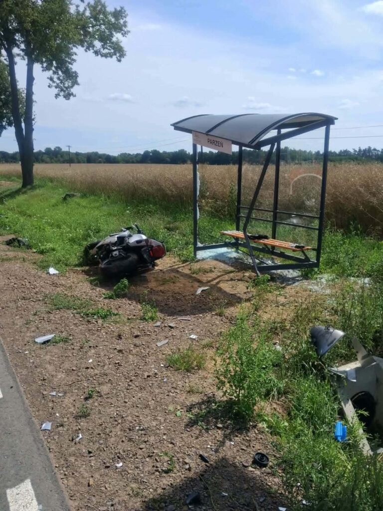 Motocykl leży przewrócony obok przystanku autobusowego wśród zieleni, z widokiem na pole.