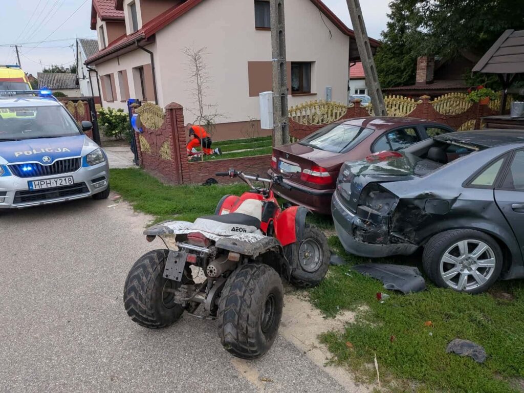Policyjny radiowóz z włączonymi sygnałami świetlnymi stoi obok uszkodzonych samochodów, a mężczyzna w pomarańczowym stroju sprząta teren.