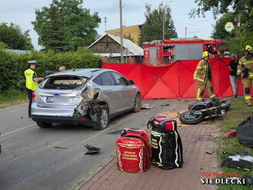 Policyjny funkcjonariusz bada zderzenie motocykla z samochodem osobowym na zablokowanej drodze, obok strażacy i sprzęt ratunkowy.