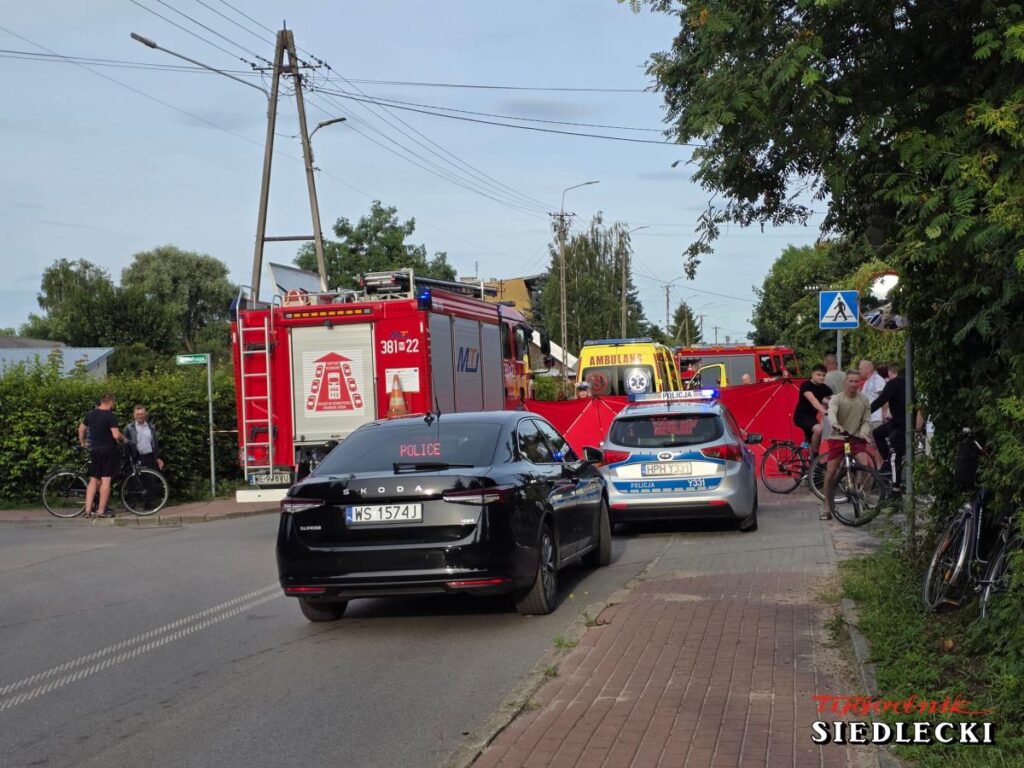Na drodze stoi radiowóz policyjny oraz samochód straży pożarnej; obok grupka osób rozmawia, a w tle widać karetki.