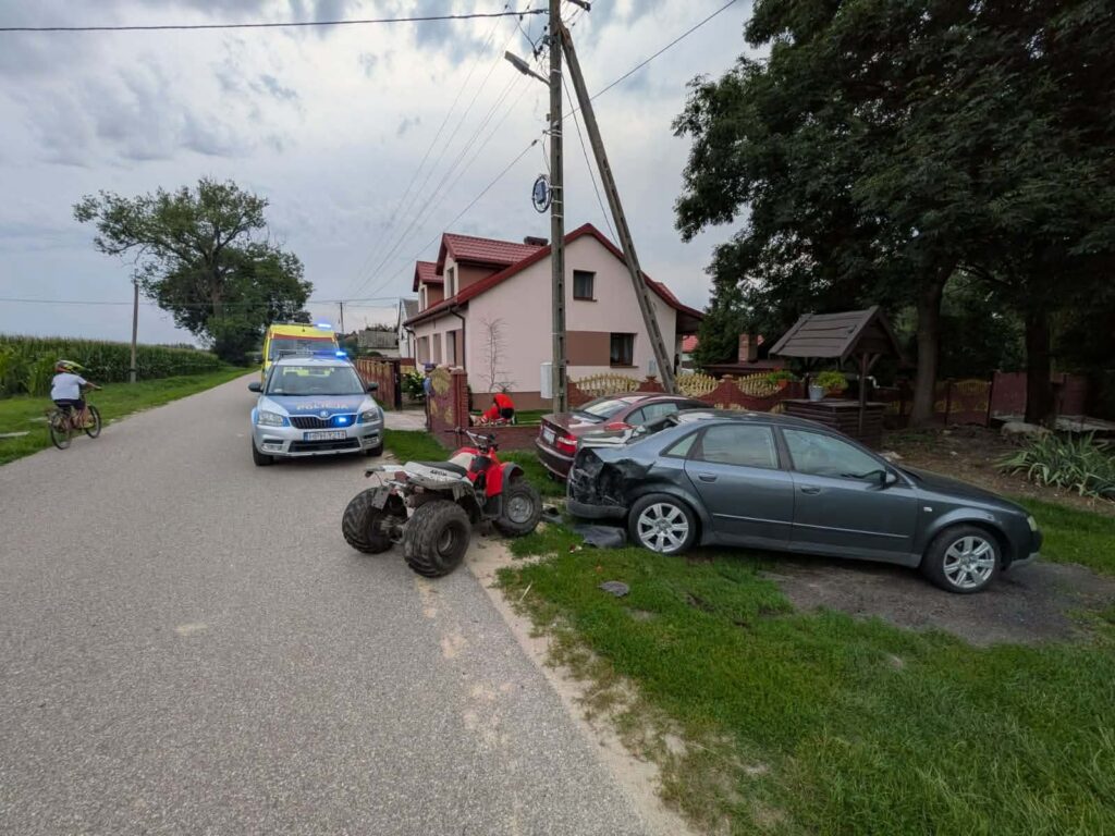 Na drodze stoi radiowóz policyjny z włączonymi sygnałami świetlnymi, obok uszkodzony samochód i quad. W tle widać dom oraz rowerzystę.