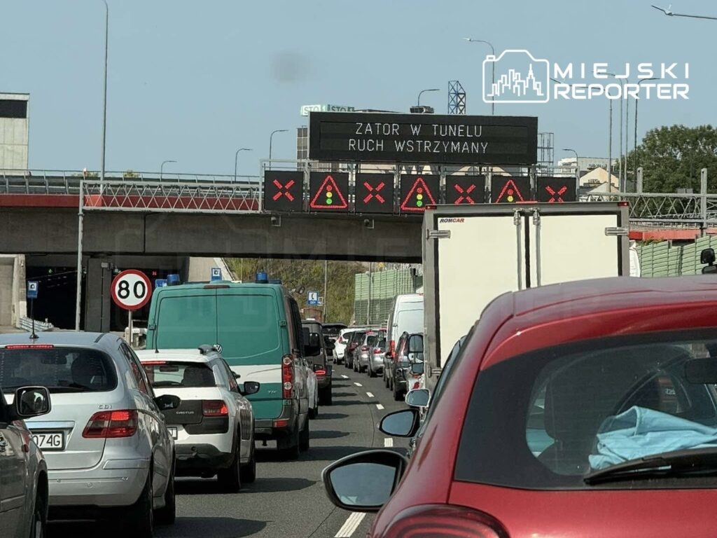 Korki w tunelu spowodowane oznakowaniem "ZATOR W TUNELU, RUCH WSTRZYMANY"; widoczne samochody osobowe i dostawcze.