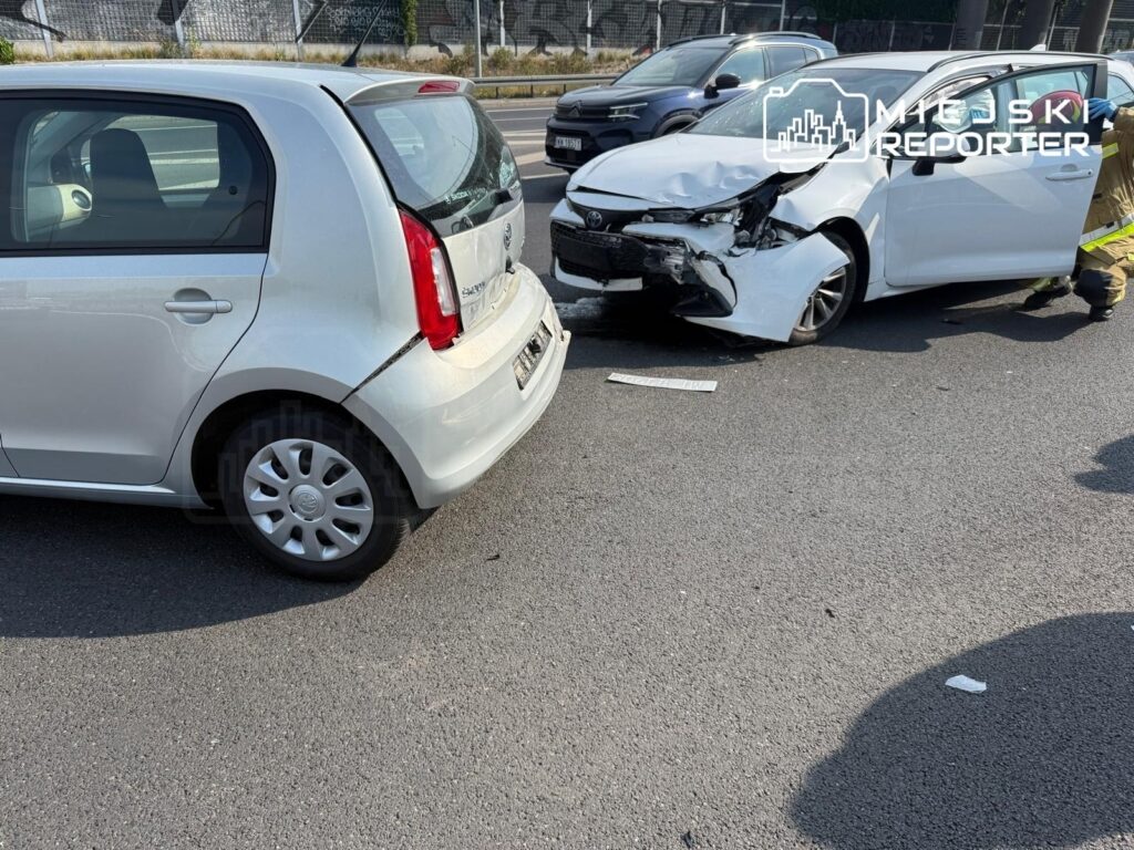 Dwa uszkodzone samochody osobowe na drodze, jeden z białym przodem, drugi z szarym tyłem, obok pracujący strażak.