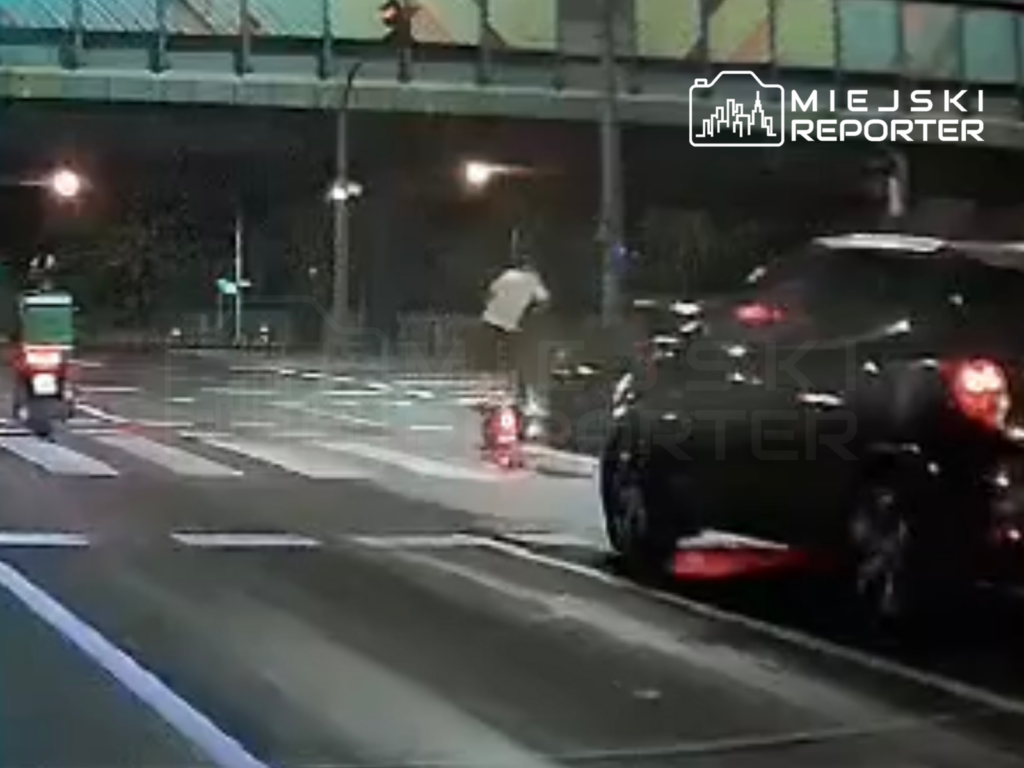 Mężczyzna na hulajnodze elektrycznej przejeżdża przez pasy na ulicy nocą, obok stojących samochodów.