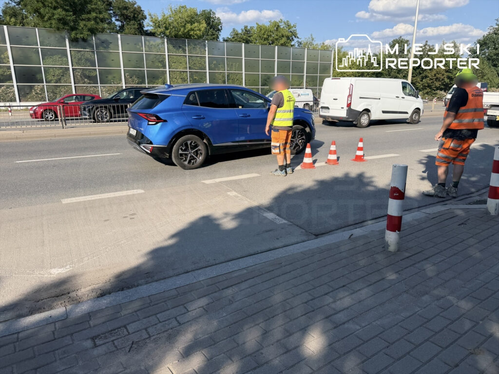 Robotnicy w kamizelkach odblaskowych kierują ruchem drogowym obok niebieskiego SUV-a na ruchliwej ulicy.