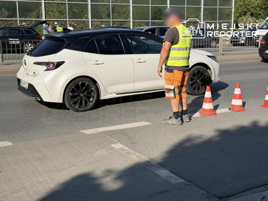 Mężczyzna w kamizelce odblaskowej kieruje ruchem drogowym obok białego samochodu osobowego na ulicy.