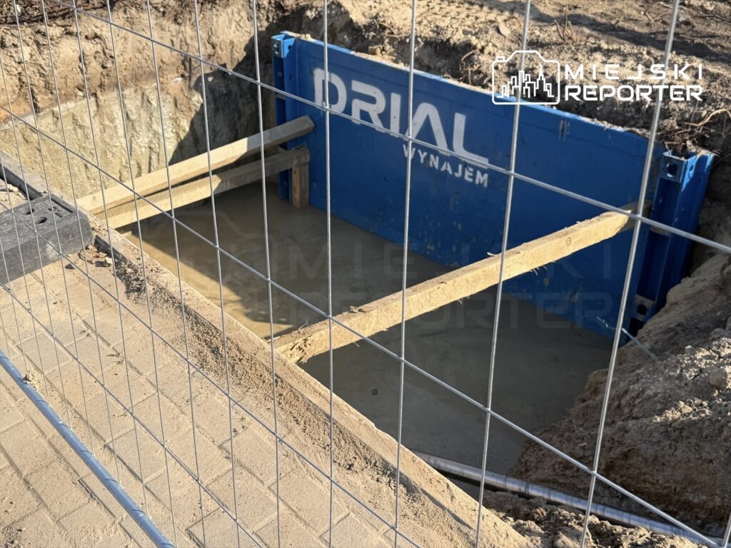 Wykop budowlany z niebieską ścianką ochronną, drewnianymi belkami i wodą na dnie, otoczony siatką.