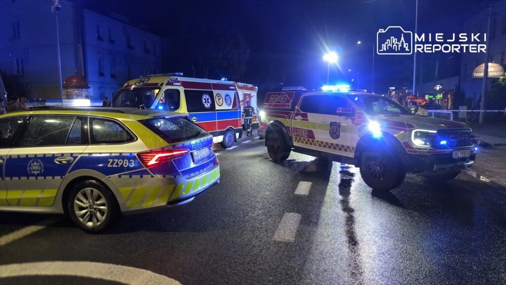 Pojazdy służb ratunkowych blokują ulicę, a ratownicy przygotowują się do akcji w nocy. W tle widać oświetlenie miasta.