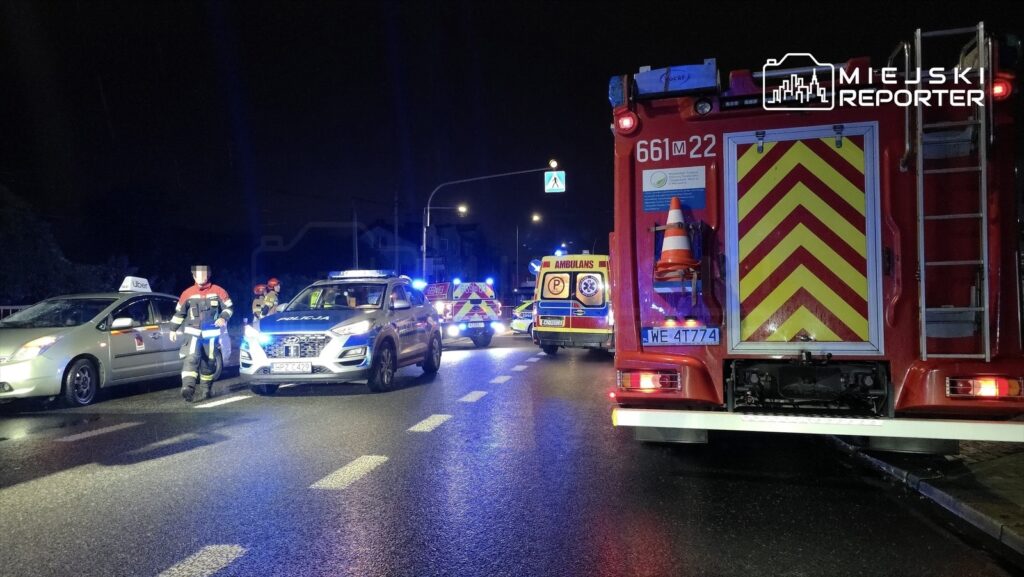 Na drodze nocą, policjant kierujący ruchem obok zaparkowanego radiowozu i wozu strażackiego, w tle karetka.