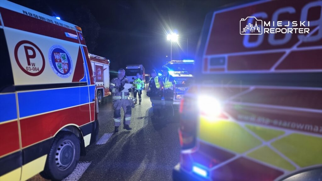 Grupa ratowników i policjantów pracuje na miejscu zdarzenia drogowego w nocy, otoczona pojazdami służb ratunkowych.