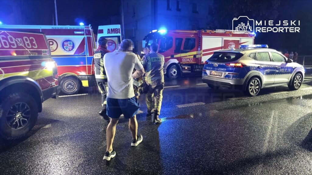 Grupa ratowników i policjantów udziela pomocy mężczyźnie w białej koszulce na mokrej jezdni w nocy.