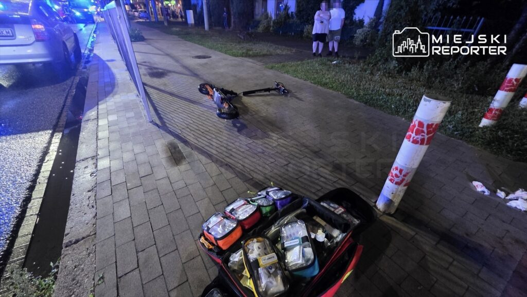 Na chodniku leży elektryczna hulajnoga, obok niej torba medyczna z wyposażeniem, w tle widać zaparkowane samochody oraz policjantów.