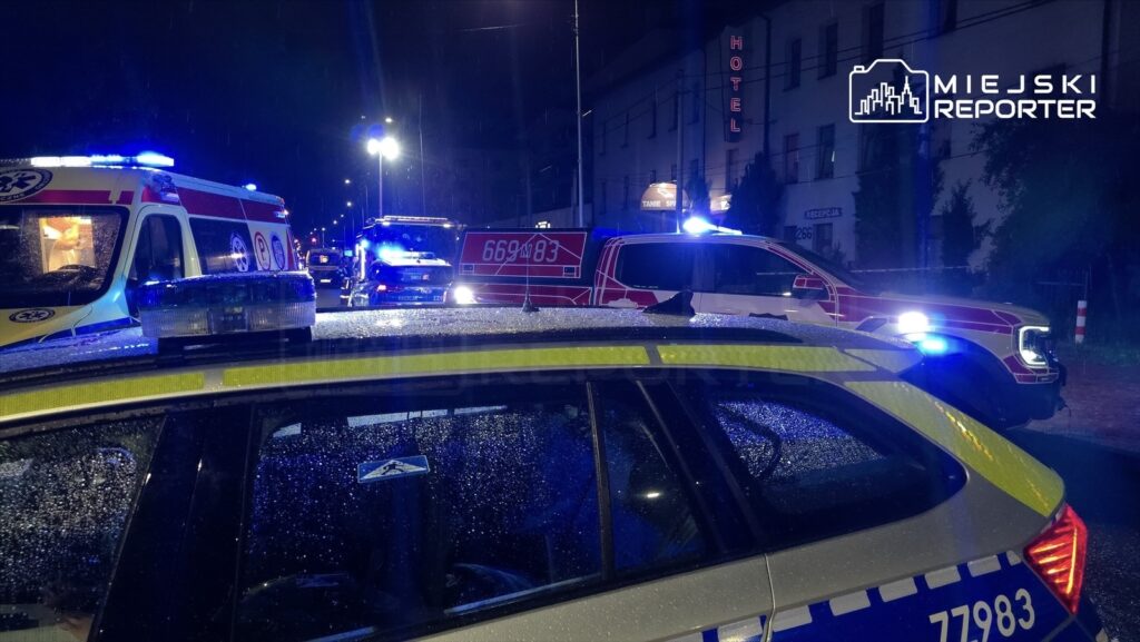 Ambulans i pojazdy policyjne z włączonymi sygnałami świetlnymi stoją na mokrej ulicy w nocy, obok hotelu.