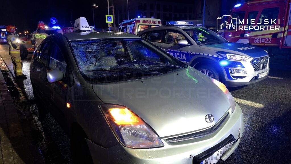 W nocy uszkodzony samochód osobowy z rozbitym przednią szybą stoi na drodze, a obok zespół ratunkowy i policja zabezpieczają miejsce zdarzenia.