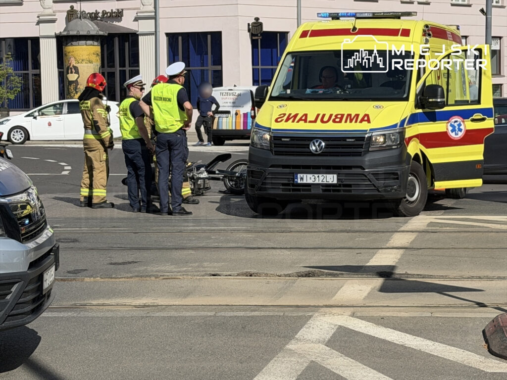 Grupa ratowników i policjantów zbliża się do przewróconego motocykla na Placu Unii, obok karetki pogotowia.