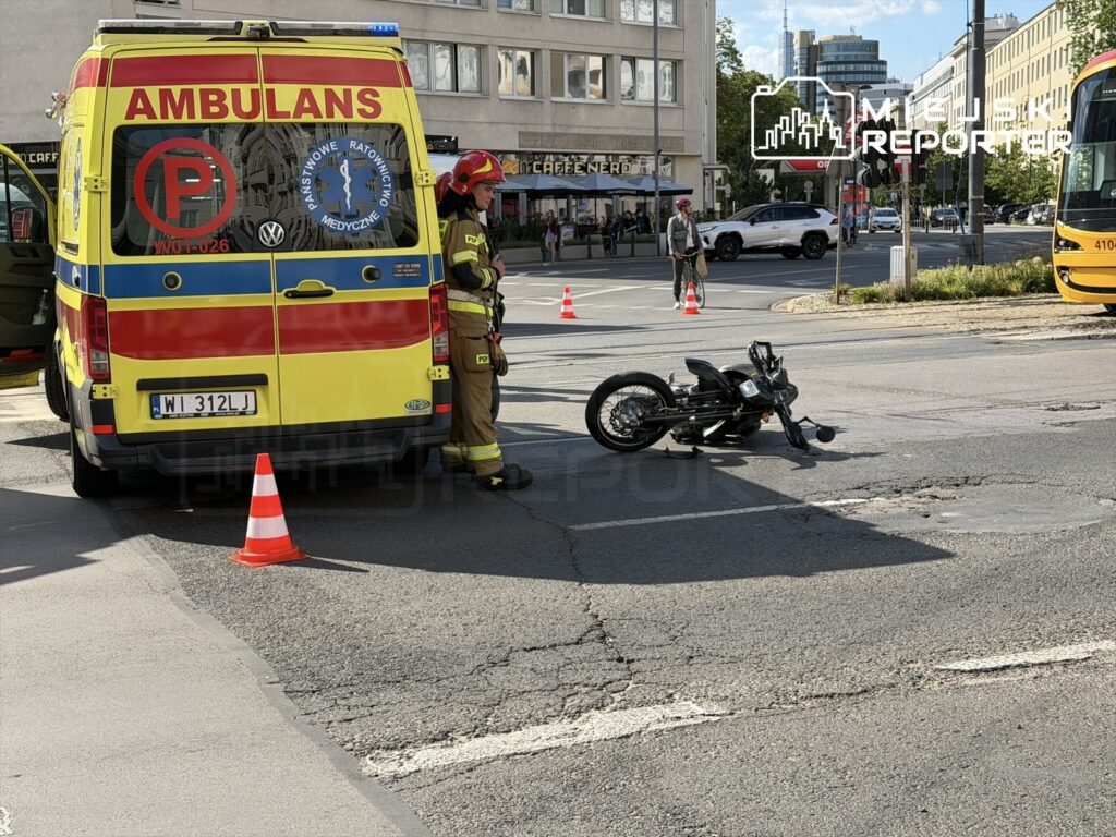 Ratownik medyczny przy ambulansie, obok leżący motocykl na jezdni. W tle ulica z samochodami i przechodniami przy Placu Unii.
