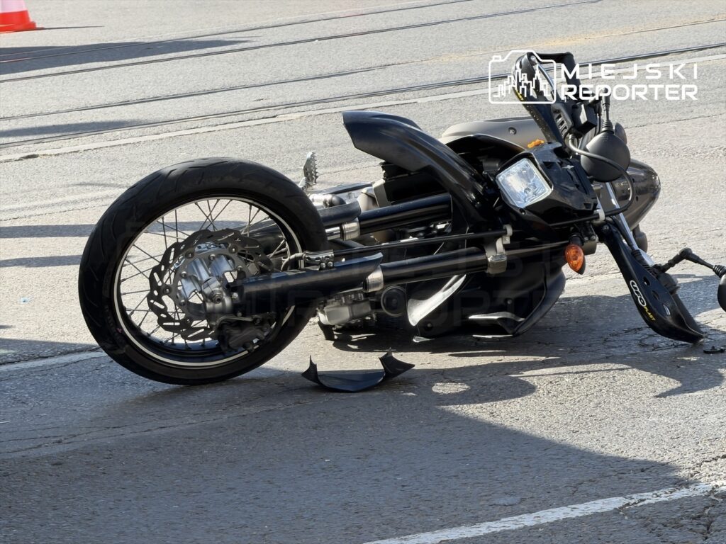 Motocykl leżący na jezdni z uszkodzoną owiewką, w otoczeniu oznaczeń drogowych na Placu Unii.