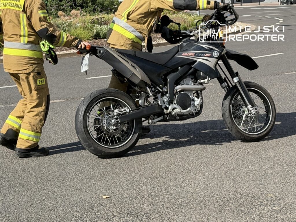 Dwaj strażacy w mundurach przekładają motocykl na jezdni, w tle widać roślinność.