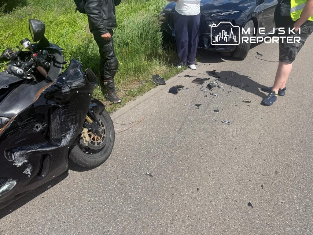 Motocykl z uszkodzoną karoserią stoi przy drodze, obok grupy osób oglądających pozostałości wypadku.