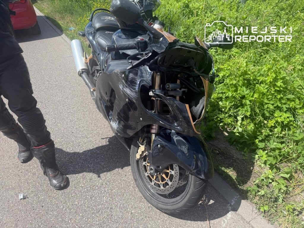 Motocyklista w skórzanej odzieży stoi obok uszkodzonego czarnego motocykla na poboczu drogi, w otoczeniu zieleni.