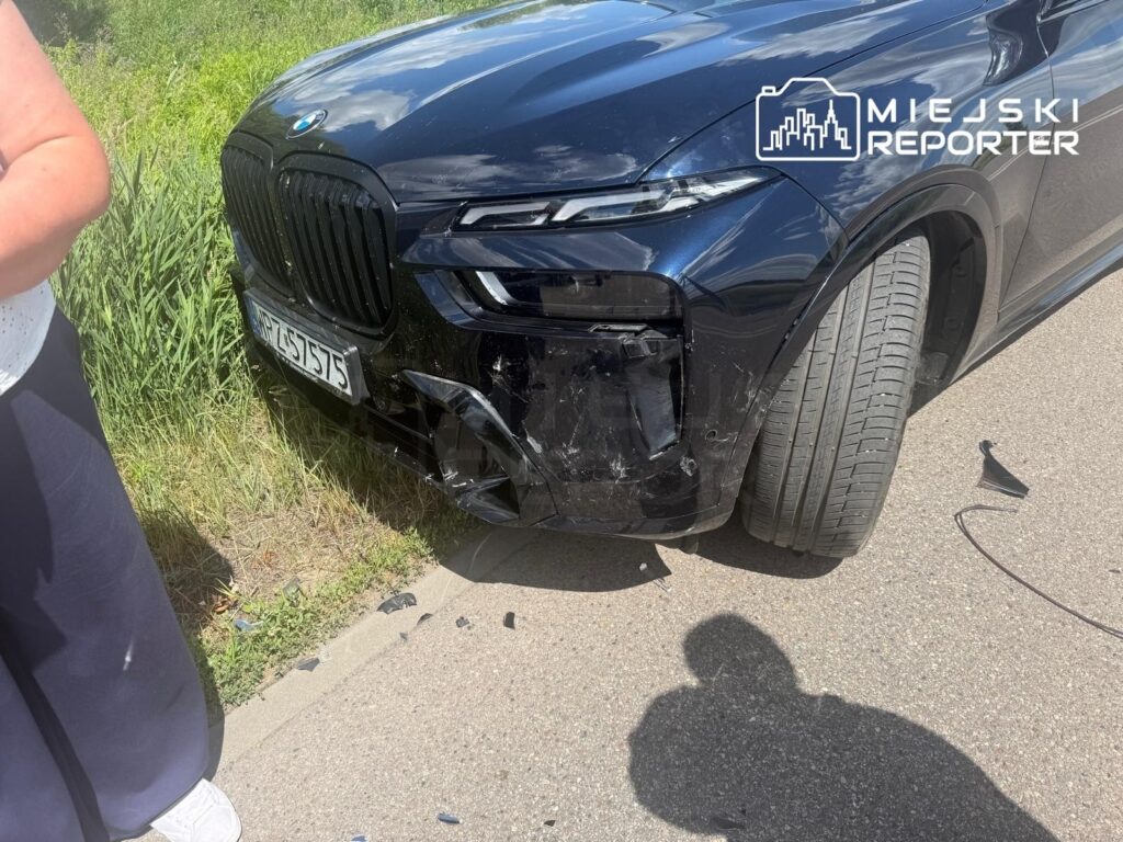 Czarny SUV z uszkodzoną przednią częścią stoi na poboczu drogi, obok widoczna osoba w białej bluzce.