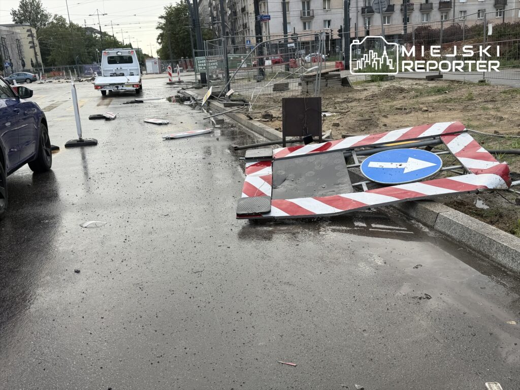 Zniszczony znak drogowy leży na mokrej nawierzchni ulicy, w otoczeniu sprzętu budowlanego i porozrzucanych elementów.