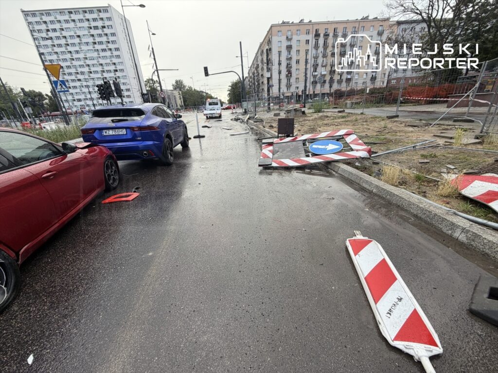 Dwa samochody stoją na mokrej jezdni, obok leżą zniszczone znaki drogowe i barierki, w tle bloki mieszkalne.