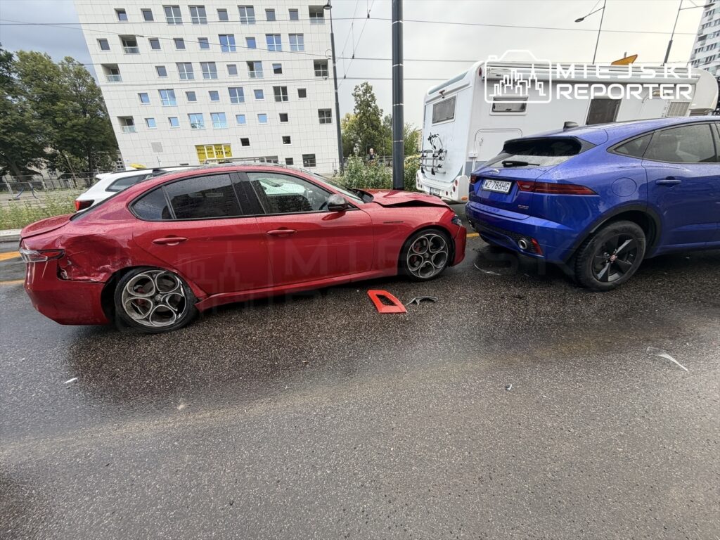 Czerwony sedan z uszkodzoną tylną częścią stoi obok niebieskiego SUV-a na deszczowej ulicy.