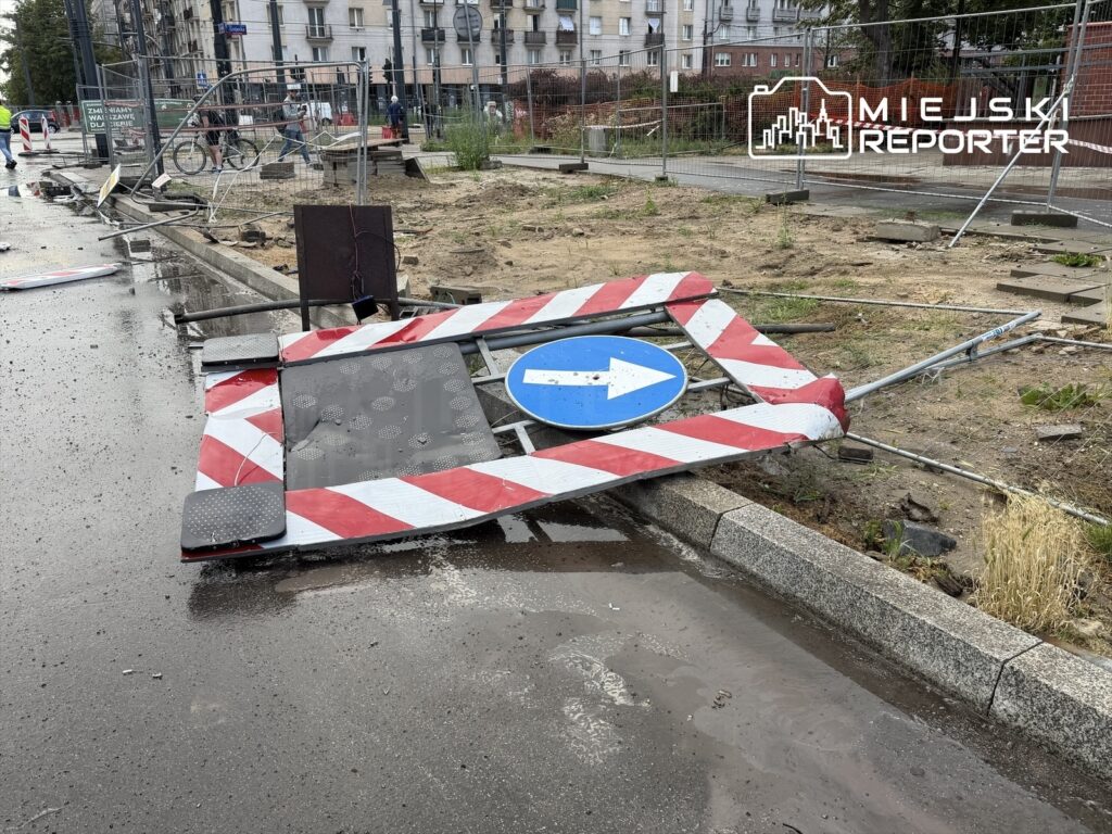 Złamany znak drogowy z niebieską strzałką leży na mokrej nawierzchni, otoczony barierkami budowlanymi.