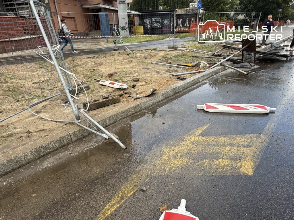 Zniszczone ogrodzenie budowlane leży na mokrej nawierzchni, a obok widać porozrzucane znaki drogowe.