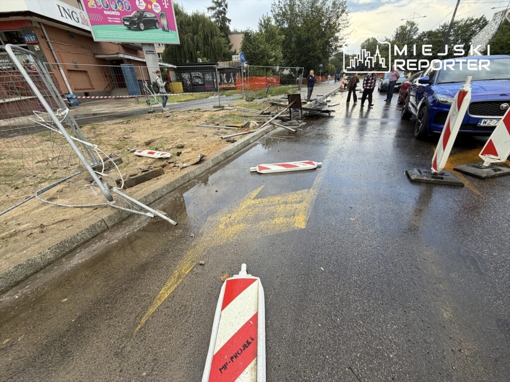 Uszkodzone ogrodzenie budowlane leży na mokrej nawierzchni drogi, obok przechodzący piesi i zaparkowane samochody.