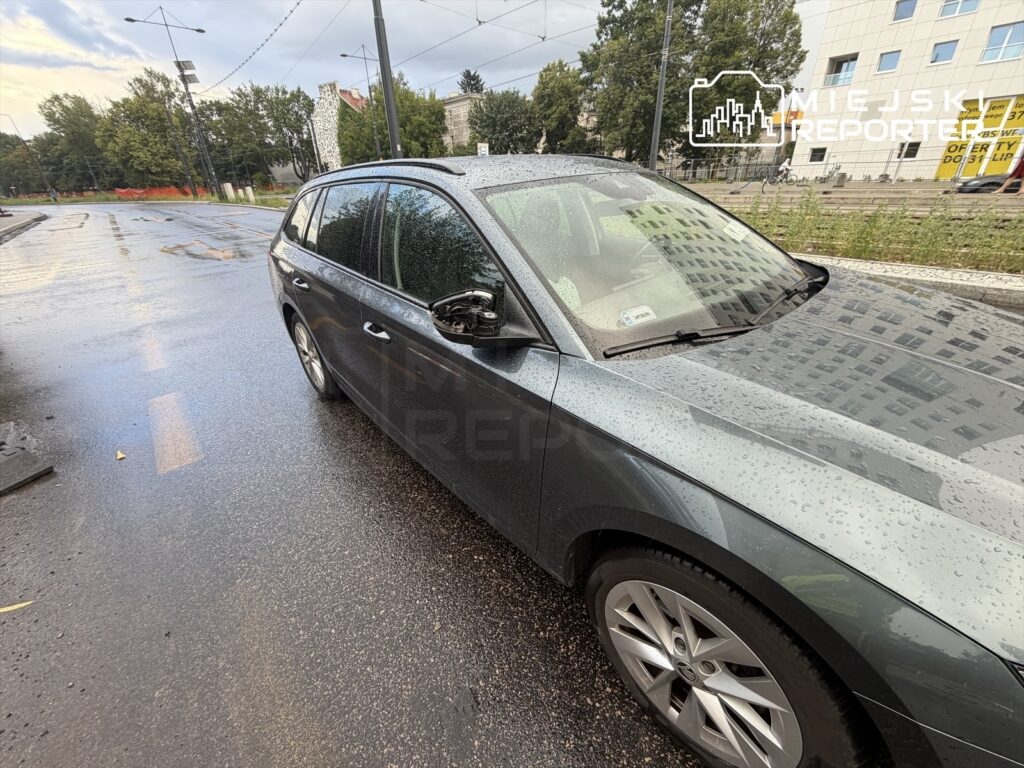 Samochód osobowy zaparkowany na mokrej jezdni, z kroplami deszczu na karoserii, w miejskim otoczeniu.