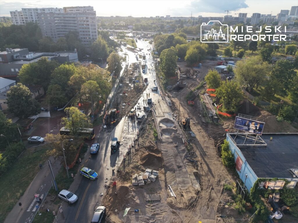 Widok na plac budowy z maszynami i pojazdami na drodze, otoczony drzewami i budynkami mieszkalnymi.