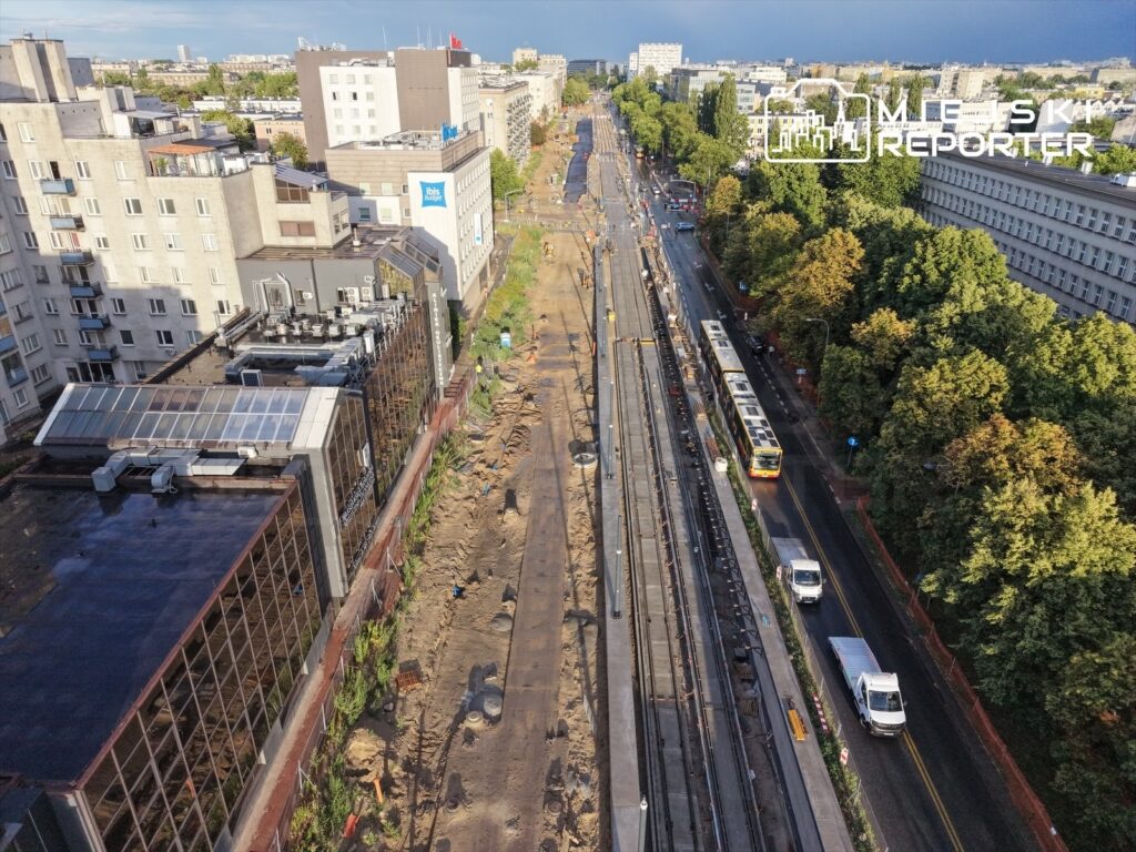 Prace budowlane na torowisku tramwajowym w miejskim otoczeniu, obok nowoczesnych budynków i drzew.