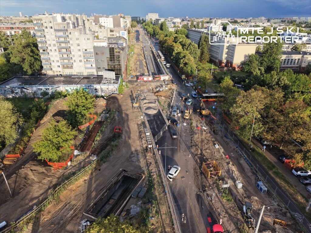 Roboty drogowe trwają na zniszczonej ulicy, z widocznymi maszynami budowlanymi i wykopami w otoczeniu zieleni.